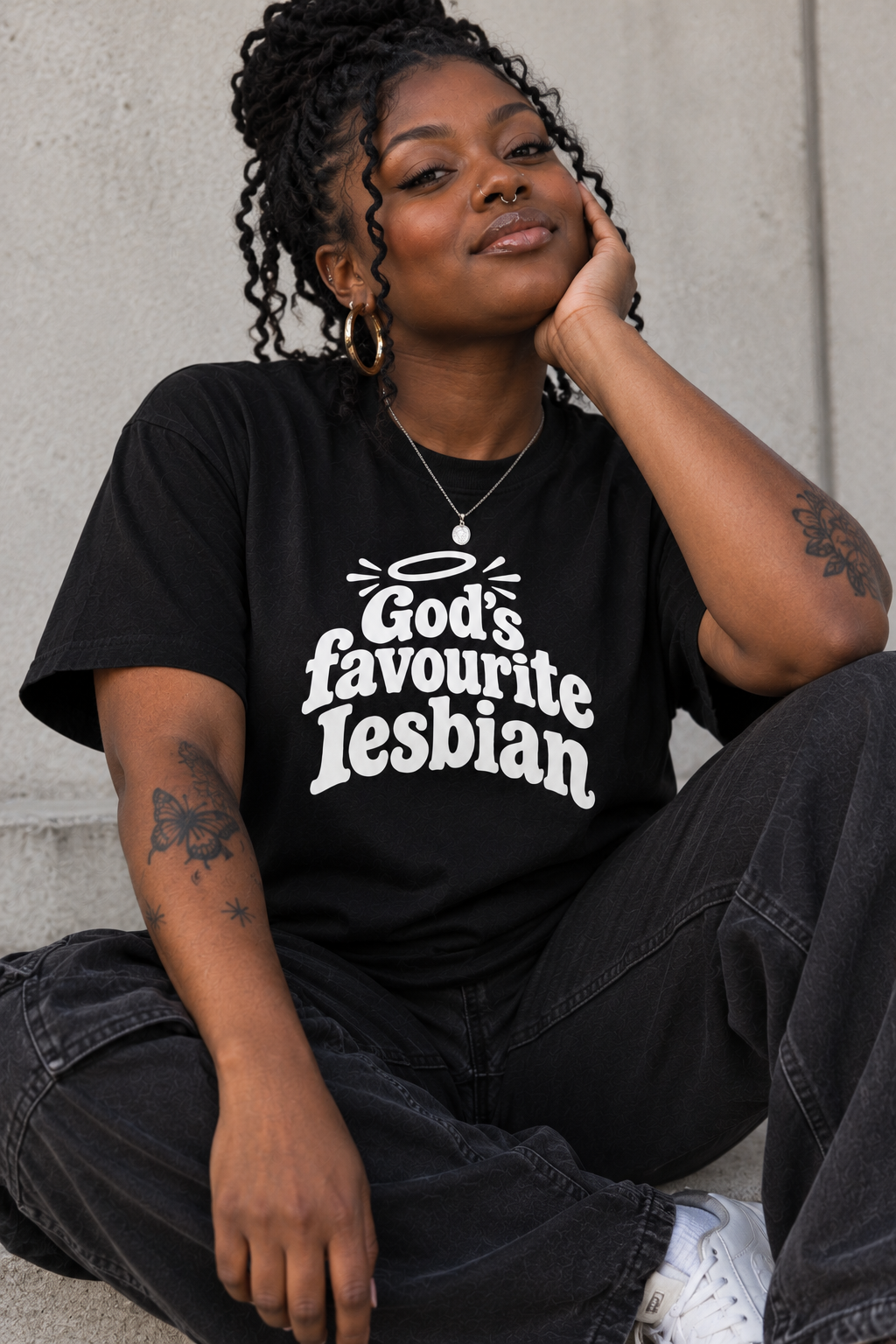 A woman sitting against a concrete wall wearing a black graphic t-shirt with the text 'God's favourite Lesbian' in white stylized lettering, paired with dark jeans and white sneakers.