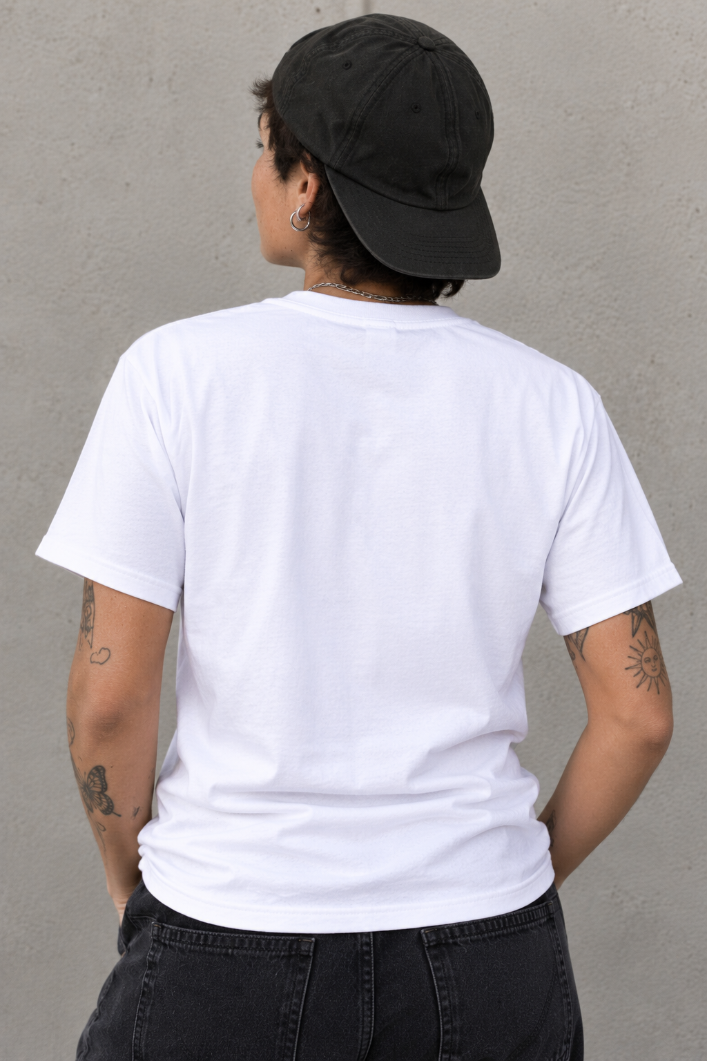 A person shown from the back wearing a plain white short-sleeve T-shirt, a black baseball cap, and dark jeans against a textured gray wall.