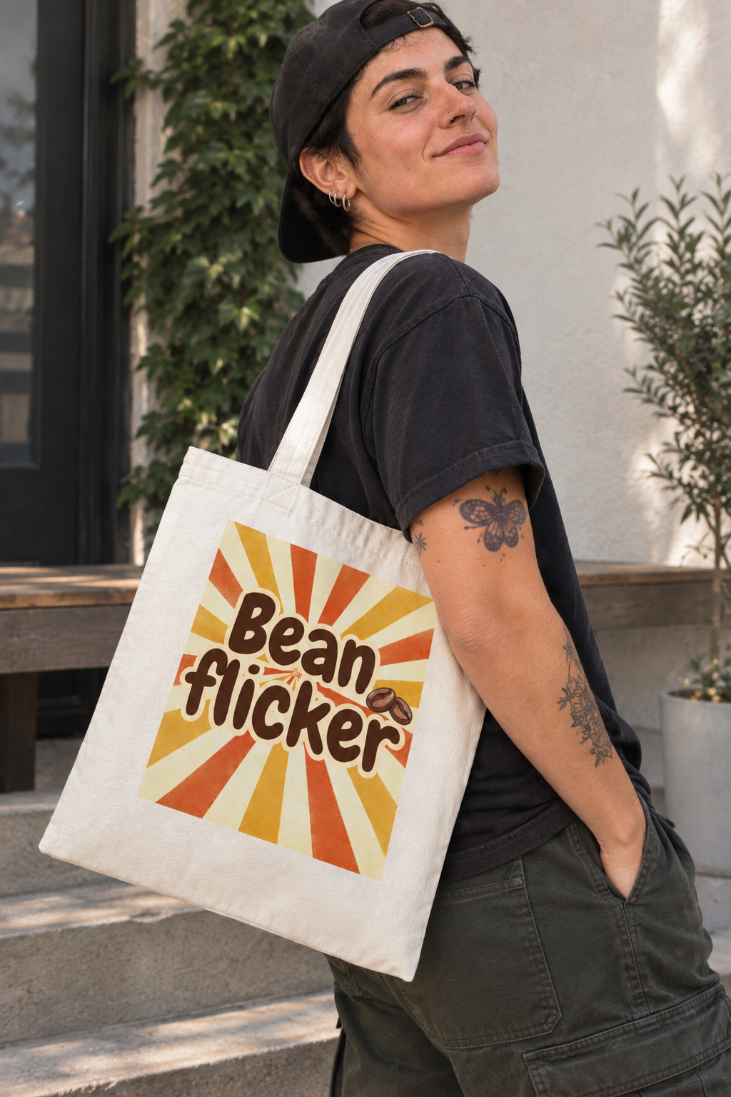 A person wearing a black cap and black t-shirt carries a cream canvas tote bag with a retro sunburst graphic that reads 'Bean flicker' in brown and orange lettering. The background shows a modern outdoor setting with a white wall, potted plants, and a wooden bench.