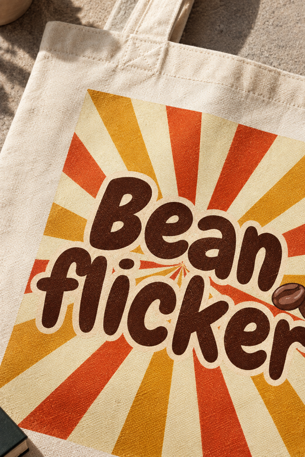 A close-up of a canvas tote bag with a retro sunburst design in orange, yellow, and cream, featuring the text 'Bean flicker' in brown with a white outline and a small coffee bean graphic.