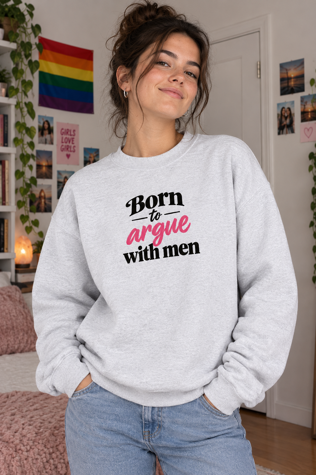 A woman wearing a light heather gray crewneck sweatshirt with the text 'Born to argue with men' printed on the front in black and pink, paired with light blue jeans in a cozy, decorated bedroom setting.