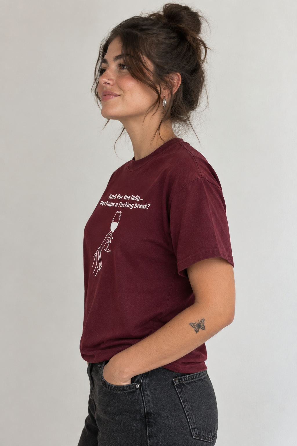A woman wearing a maroon short-sleeve graphic t-shirt and dark gray jeans, shown from a side profile against a plain light gray background.