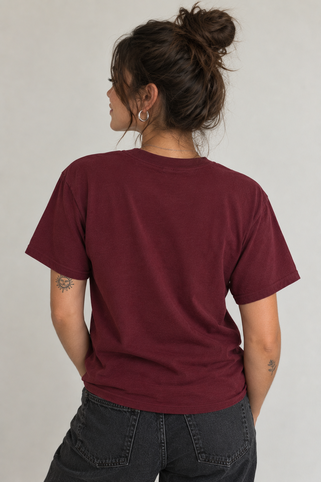 A woman shown from the back wearing a short-sleeve crewneck t-shirt in a deep burgundy color, paired with dark gray jeans, against a plain light gray background.