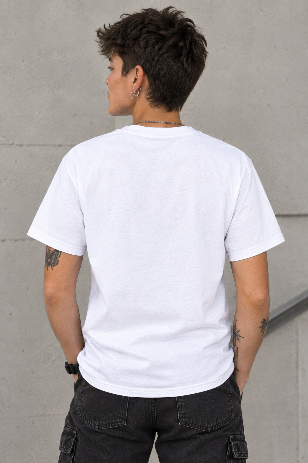 A person shown from the back wearing a plain white short-sleeve crew-neck T-shirt and dark cargo pants, standing against a light gray concrete wall.