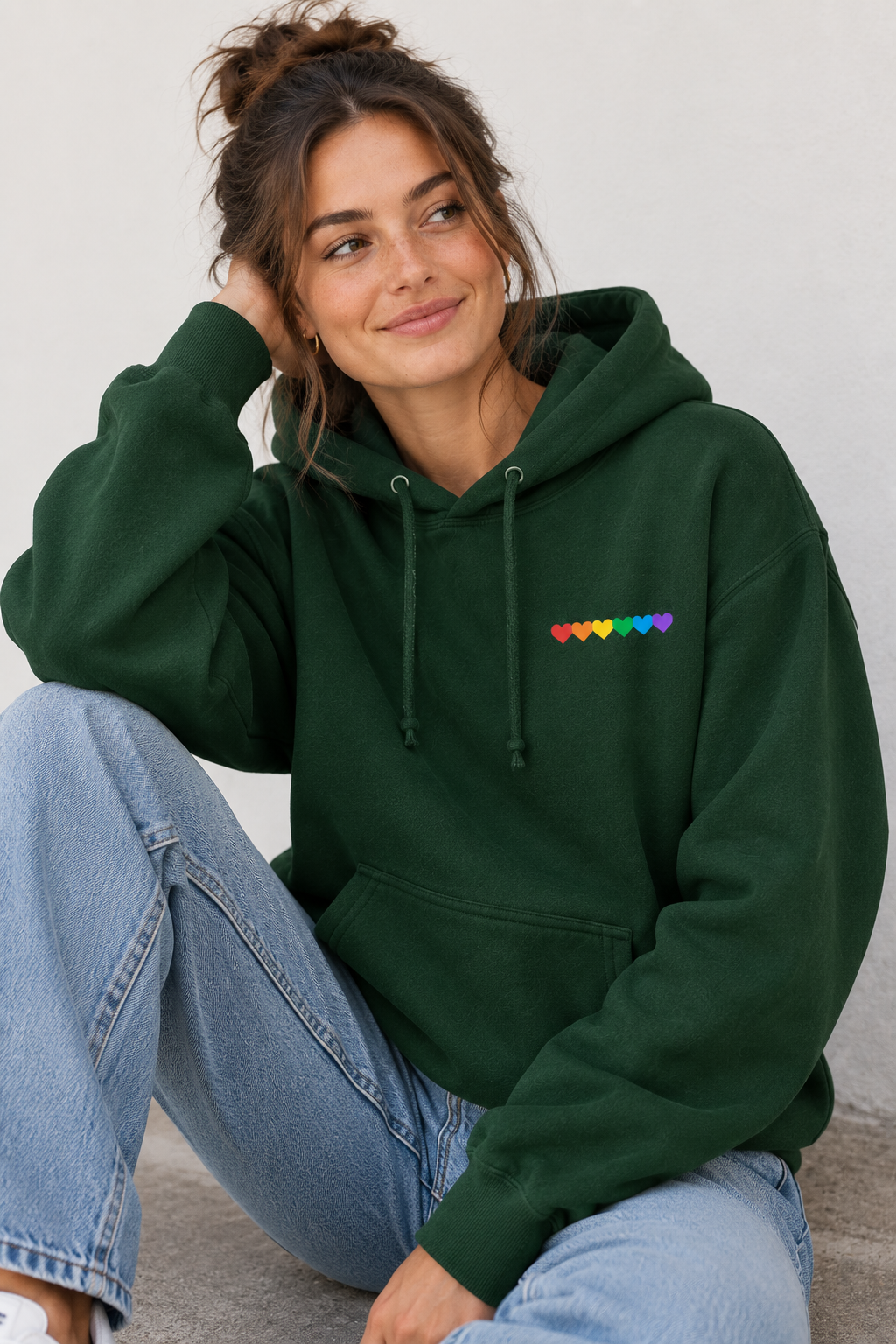 A woman wearing a dark green pullover hoodie with a small rainbow heart pattern on the left chest, paired with light blue jeans, sitting against a neutral light gray wall.