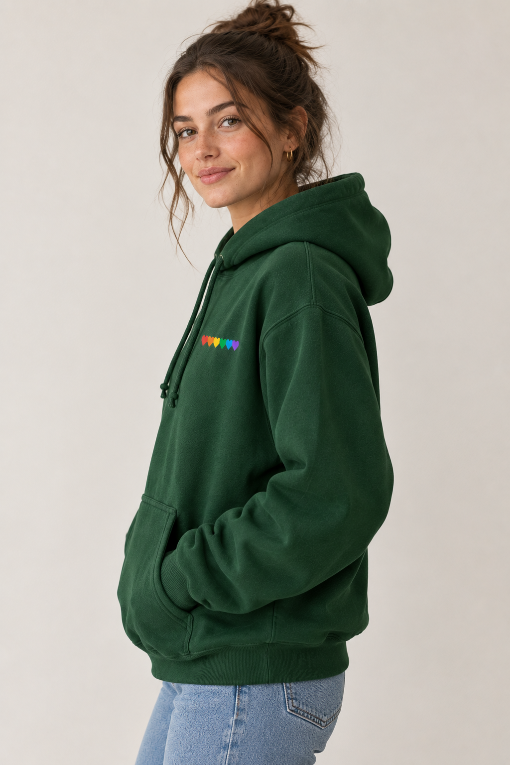 A woman wearing a dark green pullover hoodie with a small rainbow-colored 'PDAW' graphic on the left chest, paired with light blue jeans, photographed against a neutral light gray background.