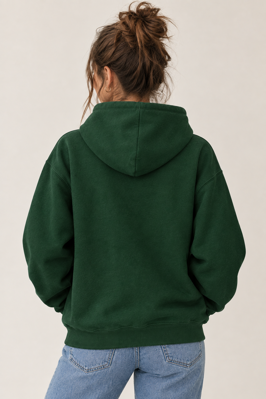 A woman viewed from behind wearing a dark forest green pullover hoodie and light blue jeans, standing against a plain light beige studio background.