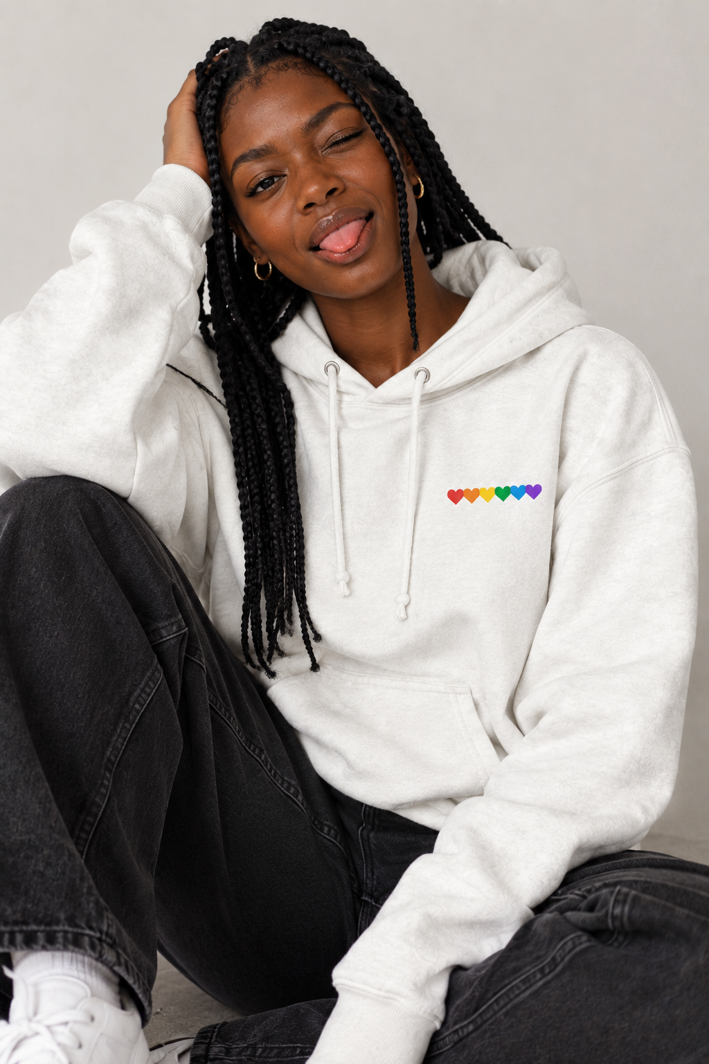 A woman with long black braids sits cross-legged wearing a white hoodie with a small rainbow heart graphic on the left chest, paired with dark gray jeans and white sneakers against a plain light gray background.