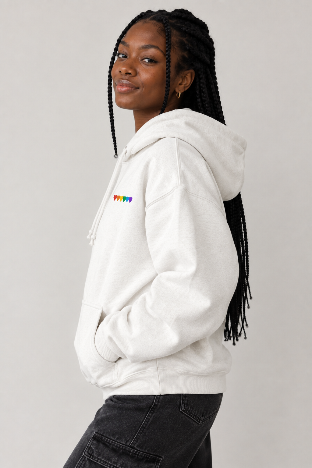 A woman with long black braids is shown from the side/back wearing a white pullover hoodie with a small rainbow-colored 'PRAWY' logo on the left chest, paired with dark gray jeans, against a plain light gray studio background.