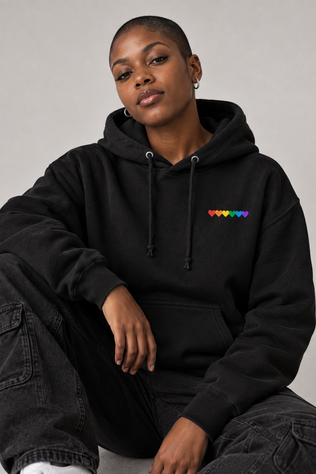 A person wearing a black pullover hoodie with a small rainbow heart graphic on the left chest, paired with dark gray cargo pants, seated against a plain light gray background.