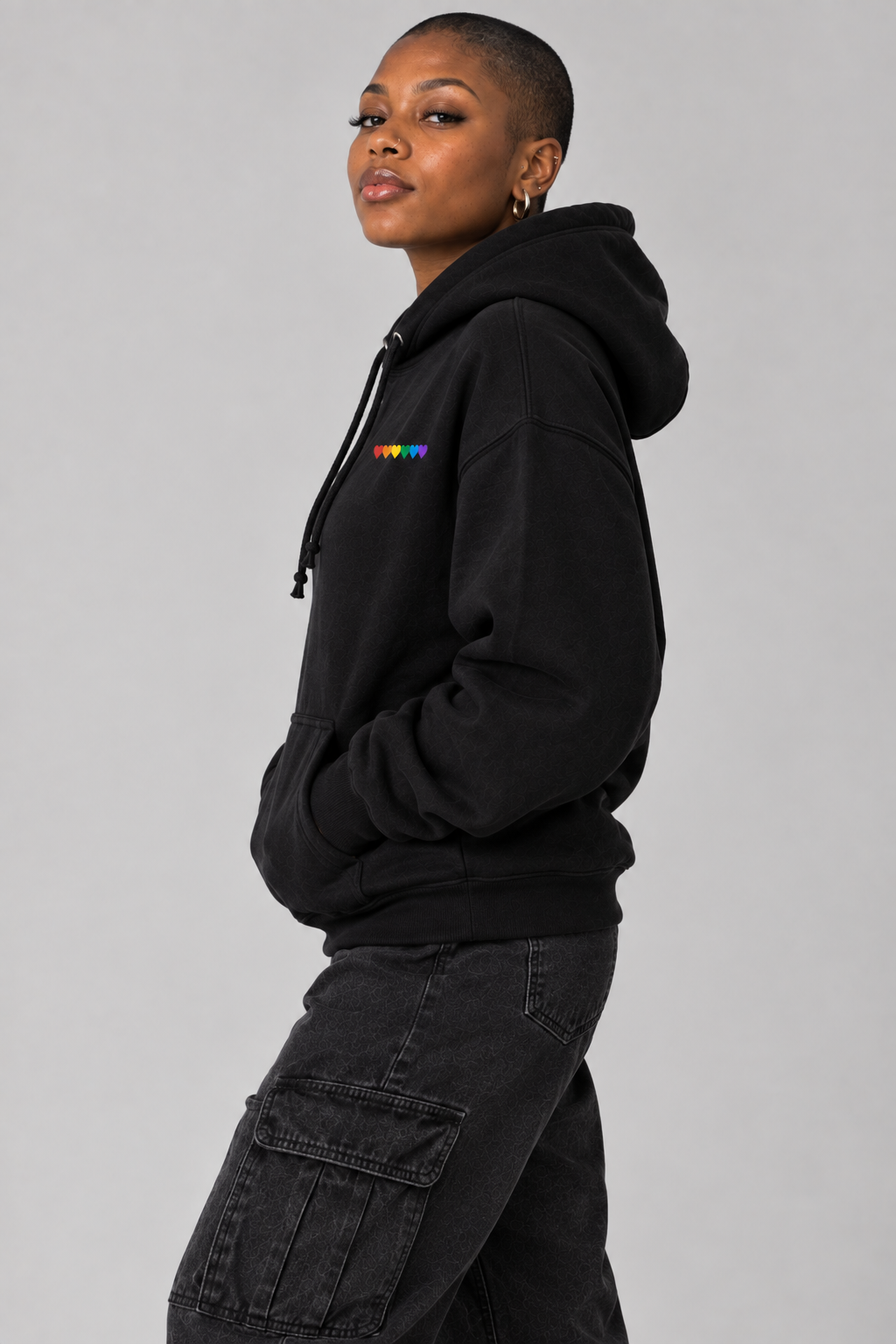 A model wearing a black pullover hoodie with a small colorful graphic on the left chest, paired with dark cargo pants, photographed against a solid gray studio background.