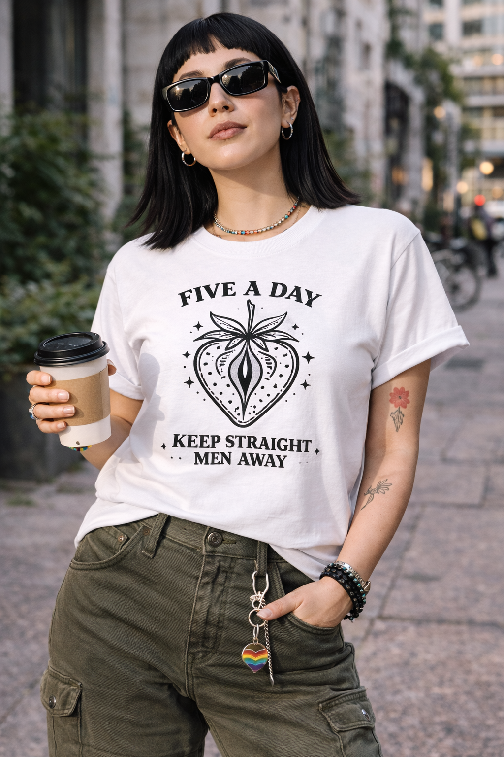 Person wearing a white t-shirt with a graphic design and text, holding a coffee cup on a city street.