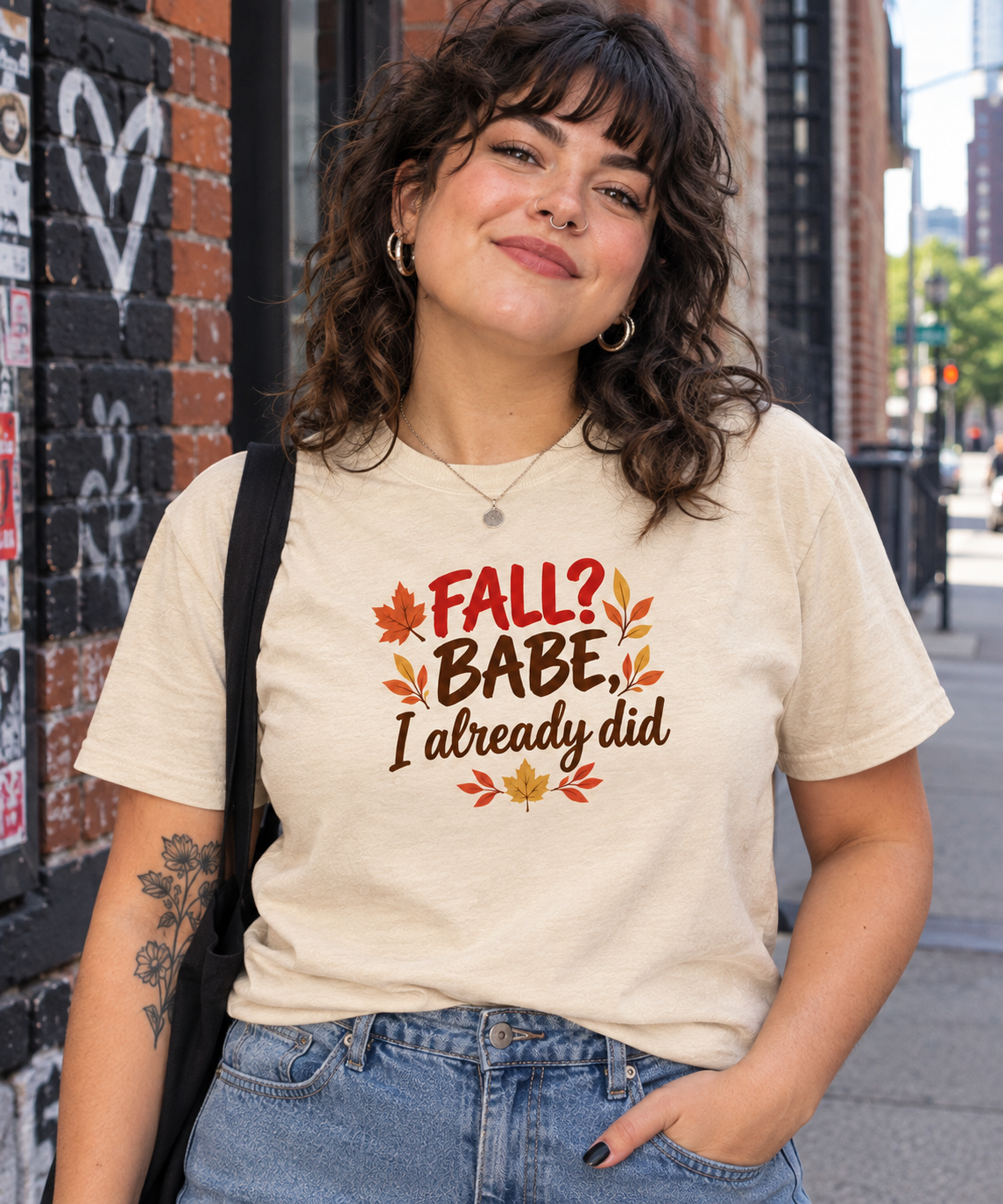 A woman wearing a beige graphic t-shirt with the text 'FALL? BABE, I already did' surrounded by autumn leaves, standing outdoors in front of a brick wall.