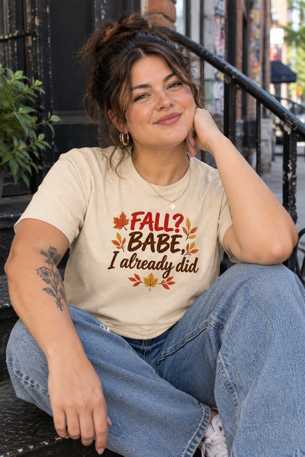 A woman sitting on outdoor steps wearing a beige graphic t-shirt with the text 'FALL? BABE, I already did' in brown and orange lettering with autumn leaves.