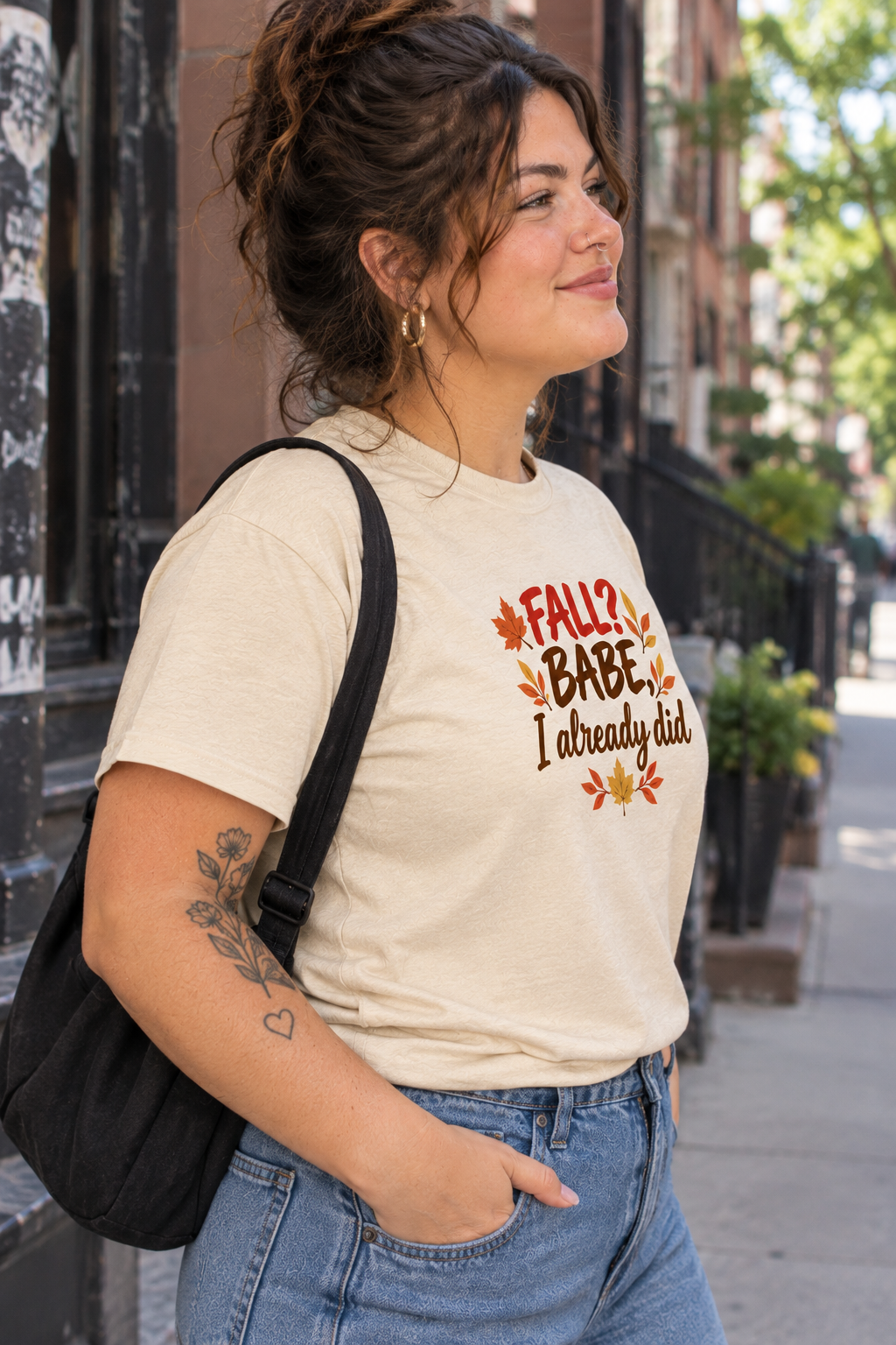 A woman wearing a cream-colored graphic t-shirt with a fall-themed design that reads 'FALL? BABE I already did' surrounded by orange and brown leaves. She is carrying a black shoulder bag and wearing blue jeans, standing on an outdoor city sidewalk.