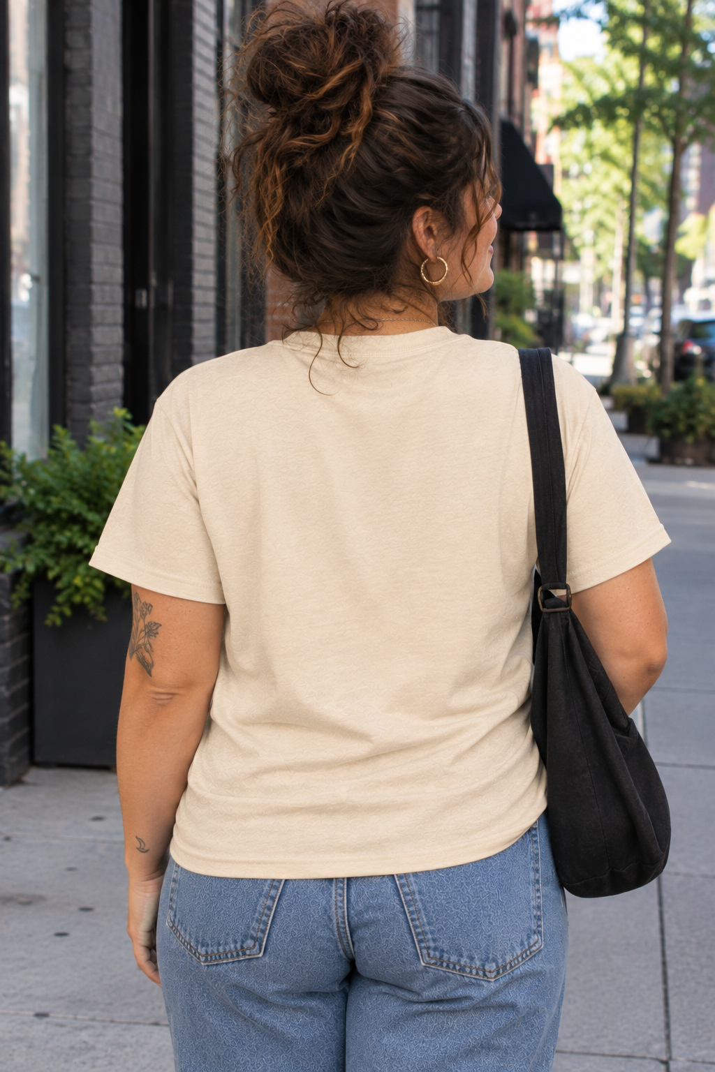 A woman viewed from behind wearing a light beige short-sleeve t-shirt and blue jeans, carrying a black shoulder bag on a city sidewalk.