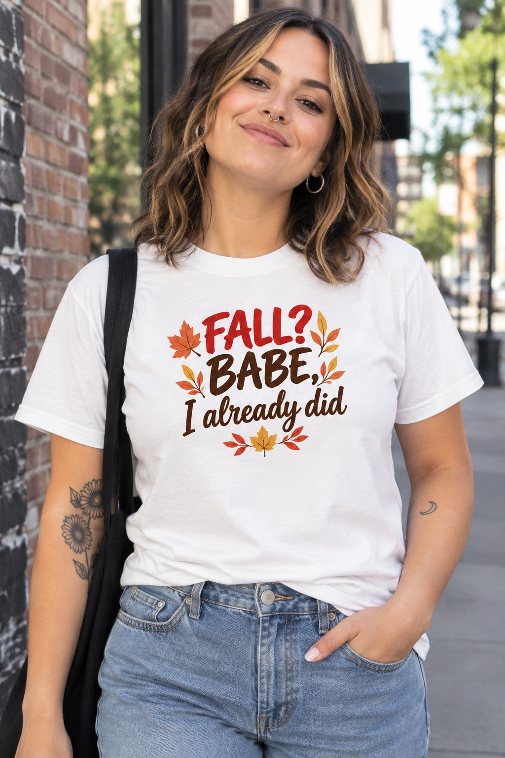 A woman wearing a white graphic t-shirt with the text 'FALL? BABE, I already did' and autumn leaf illustrations, paired with blue jeans, standing outdoors next to a brick wall.