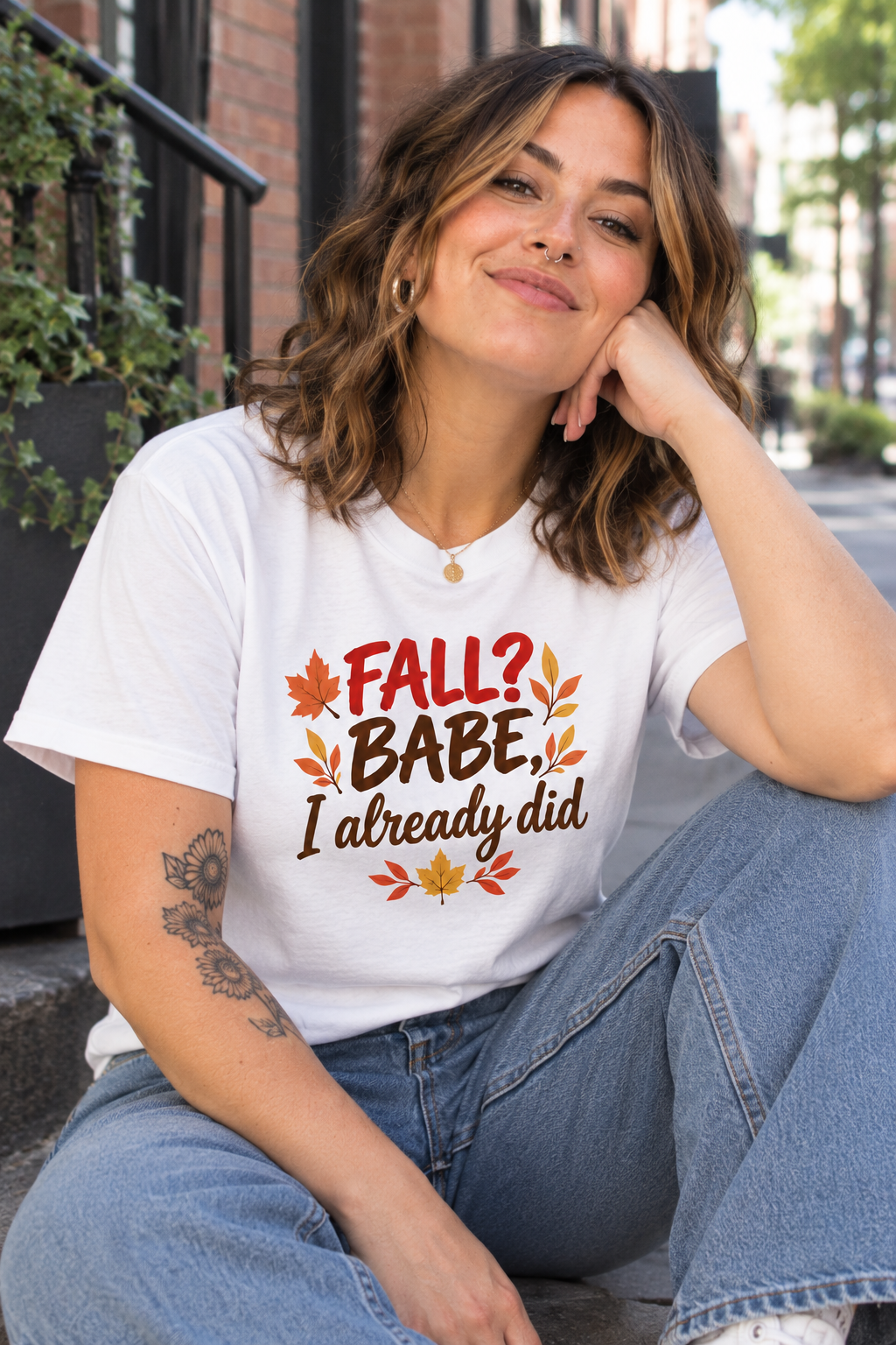 A woman sitting outdoors on a city street wearing a white graphic T-shirt that reads 'FALL? BABE, I already did' with autumn leaves, paired with light blue jeans.