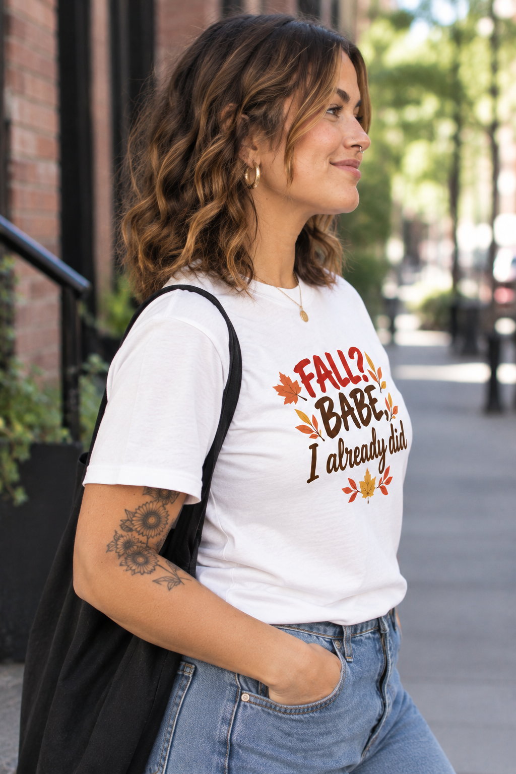A woman wearing a white graphic t-shirt with the text 'FALL 5 BABE I already did' in red and brown lettering with autumn leaves, paired with blue jeans and a black tote bag, standing on an outdoor city sidewalk.