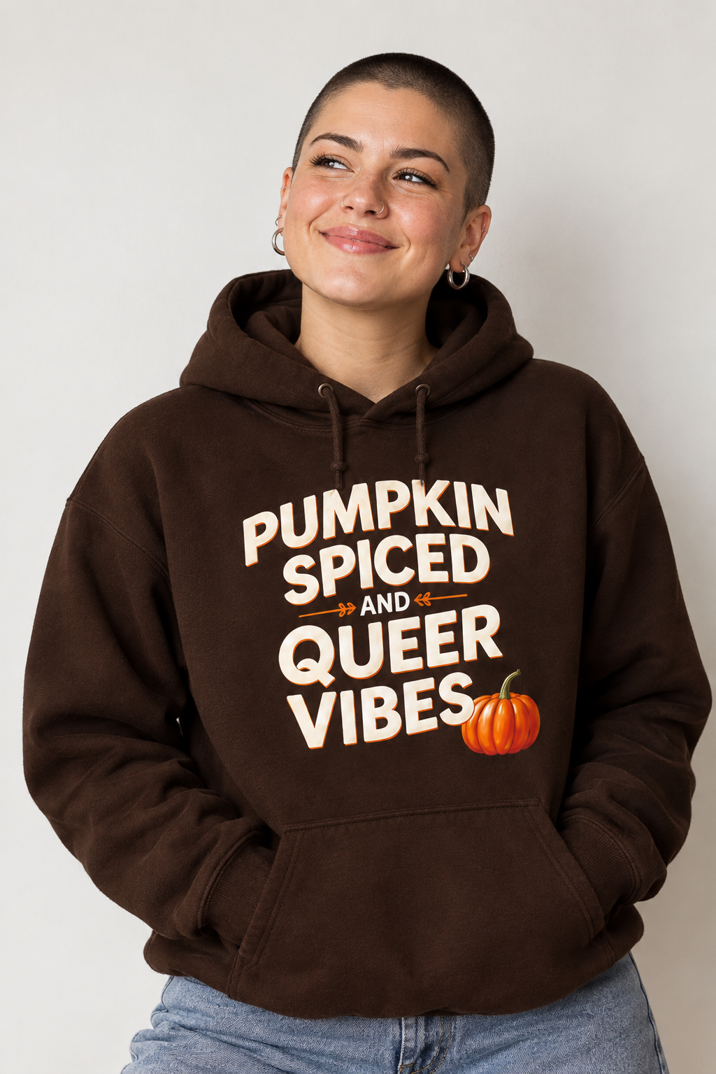 A woman with a shaved head wearing a dark brown pullover hoodie with the text 'PUMPKIN SPICED AND QUEER VIBES' in cream lettering, accented with an orange pumpkin graphic, against a plain light gray background.