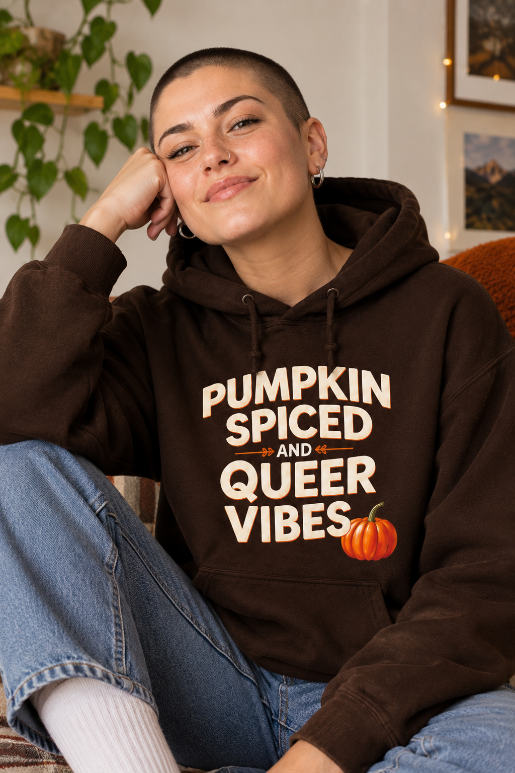 A woman wearing a dark brown hoodie with the text 'PUMPKIN SPICED AND QUEER VIBES' in cream lettering, sitting in a cozy indoor setting.