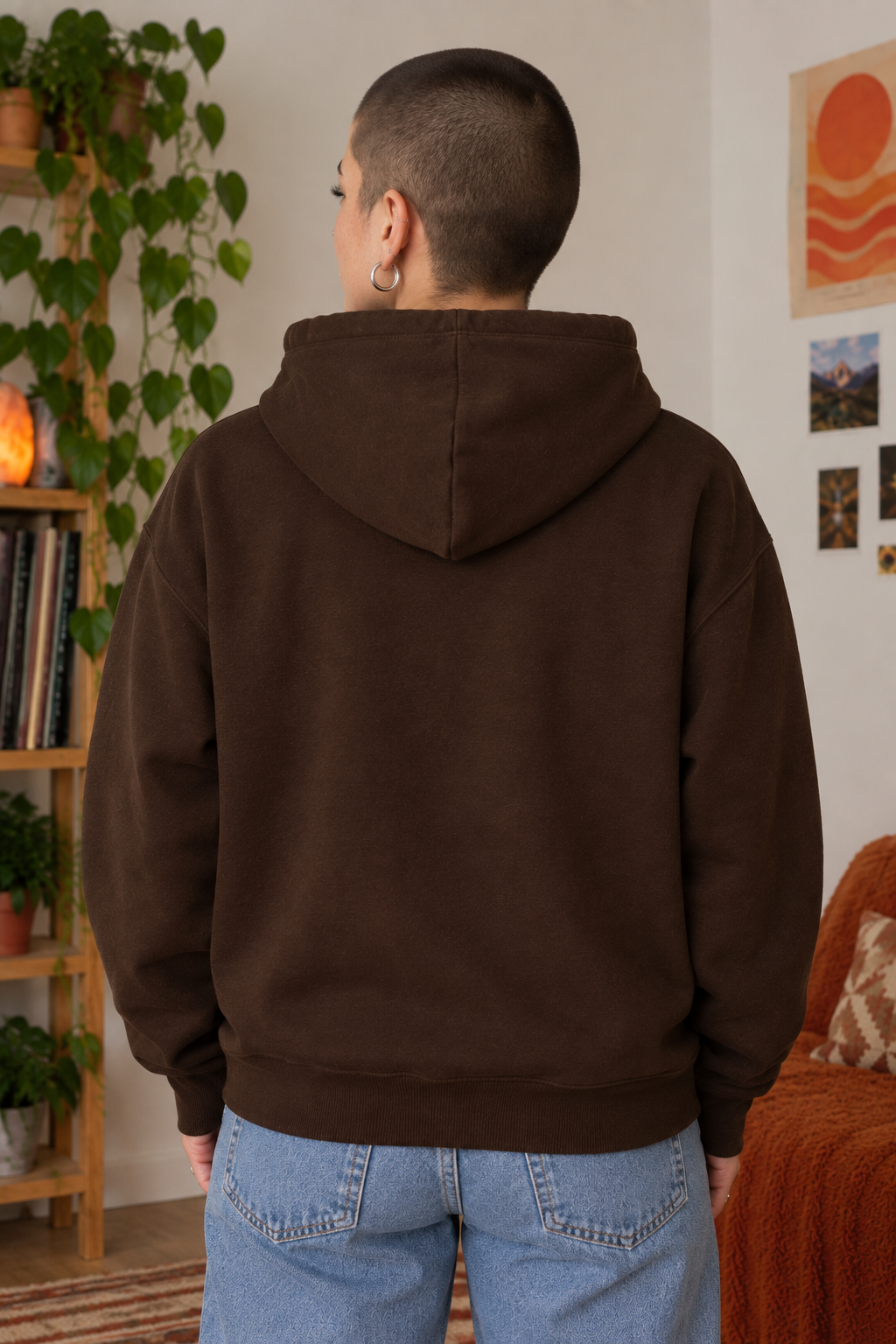 A person viewed from behind wearing a dark brown pullover hoodie and light blue jeans in a cozy indoor room with plants, a bookshelf, and wall art.
