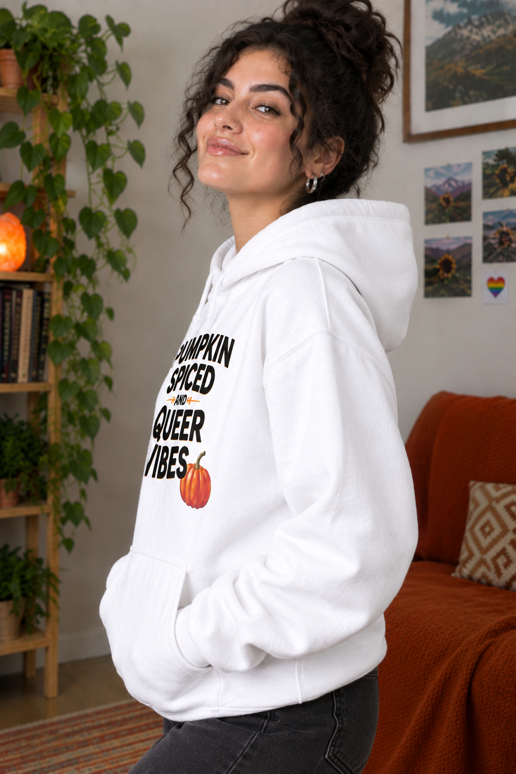 A woman wearing a white pullover hoodie with the text 'PUMPKIN SPICED AND QUEER VIBES' in black, accented by small orange arrows and a small orange pumpkin graphic. She is posing in a cozy, warmly lit room with a bookshelf, plants, and a couch in the background.