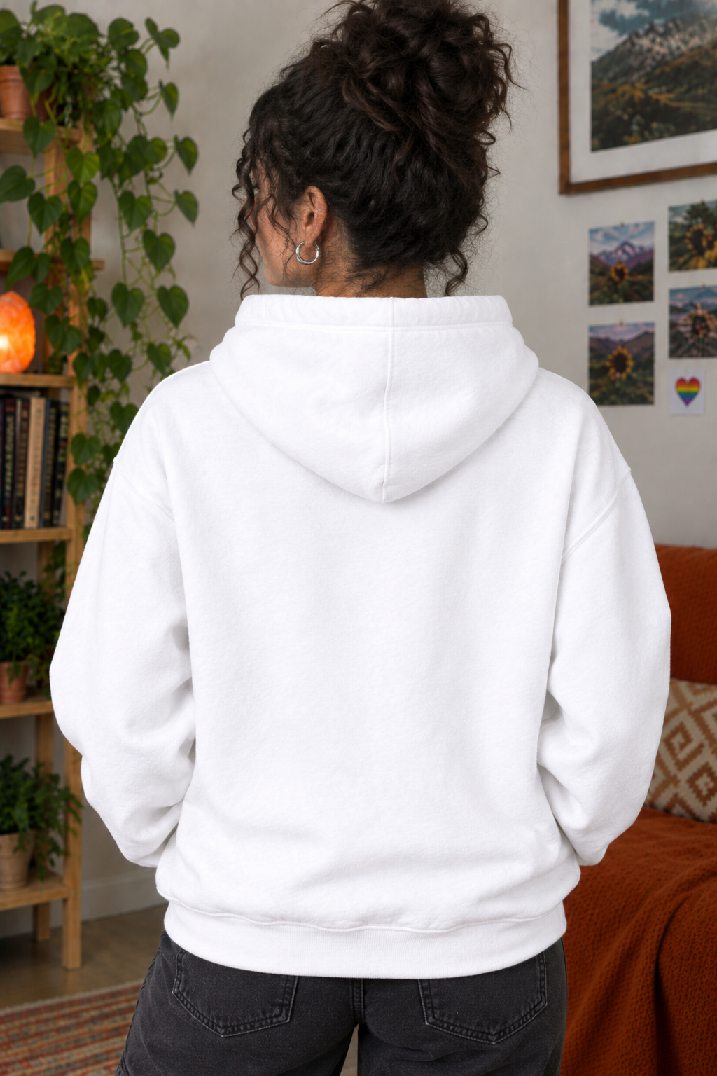 A person shown from behind wearing a plain white pullover hoodie in a cozy indoor room with plants, framed wall art, and an orange sofa.