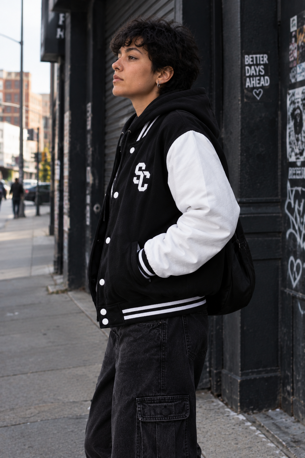 A person standing outdoors on a city sidewalk wearing a black and white varsity-style jacket over a black hoodie and black cargo pants. The jacket has white sleeves, white ribbed trim, and a white embroidered 'SC' logo on the left chest.