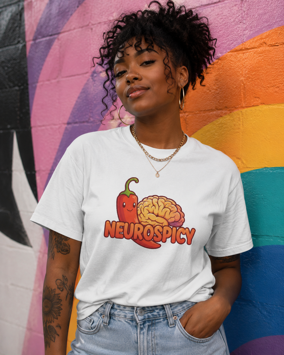 A woman wearing a white graphic t-shirt with a 'Neurospicy' design featuring a red chili pepper and a brain, standing in front of a colorful painted brick wall.