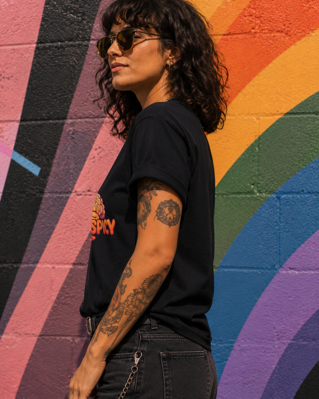 A woman with dark curly hair and sunglasses, wearing a black graphic t-shirt and dark jeans, standing in profile against a colorful rainbow mural painted on a brick wall.