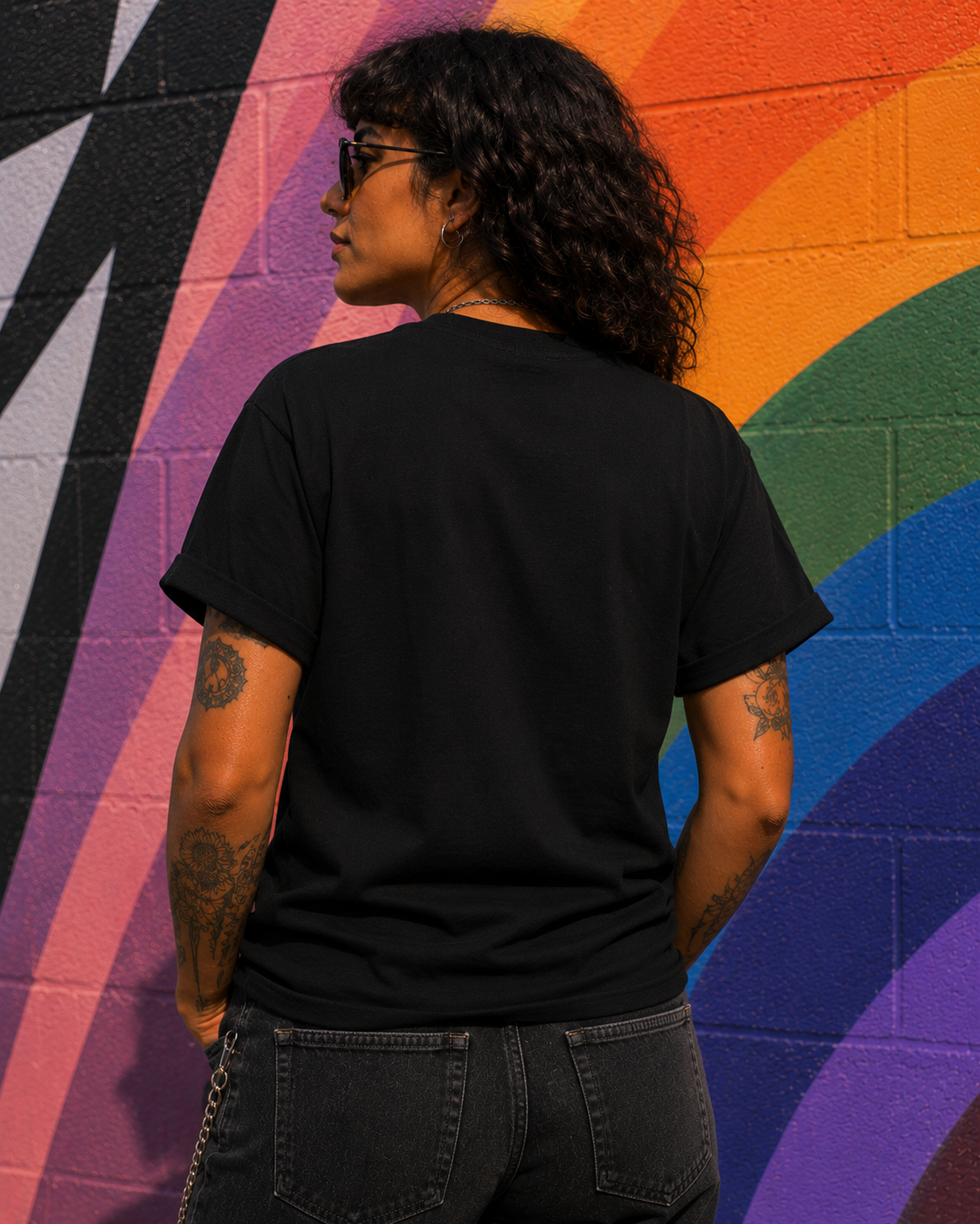 A person with curly dark hair and tattoos, seen from behind, wearing a plain black t-shirt and dark jeans, standing in front of a vibrant rainbow-colored brick wall.