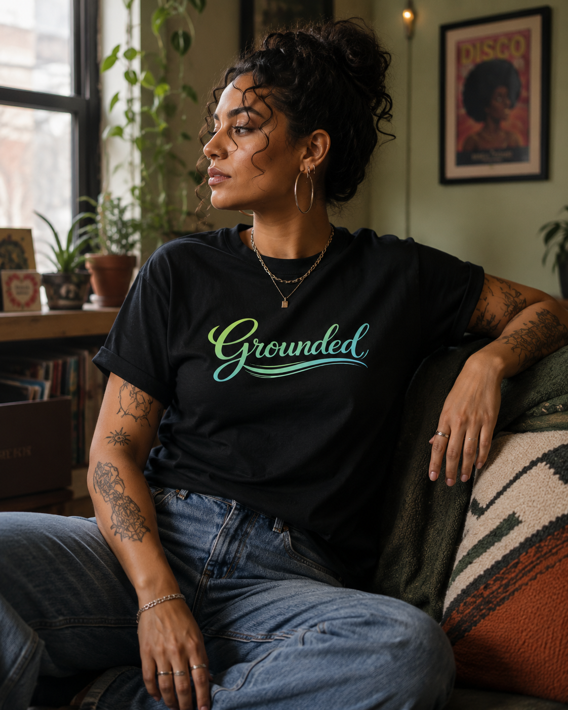 A woman sitting on a couch in a cozy room, wearing a black t-shirt with the word 'Grounded' printed in a teal and green script font. She is also wearing blue jeans.