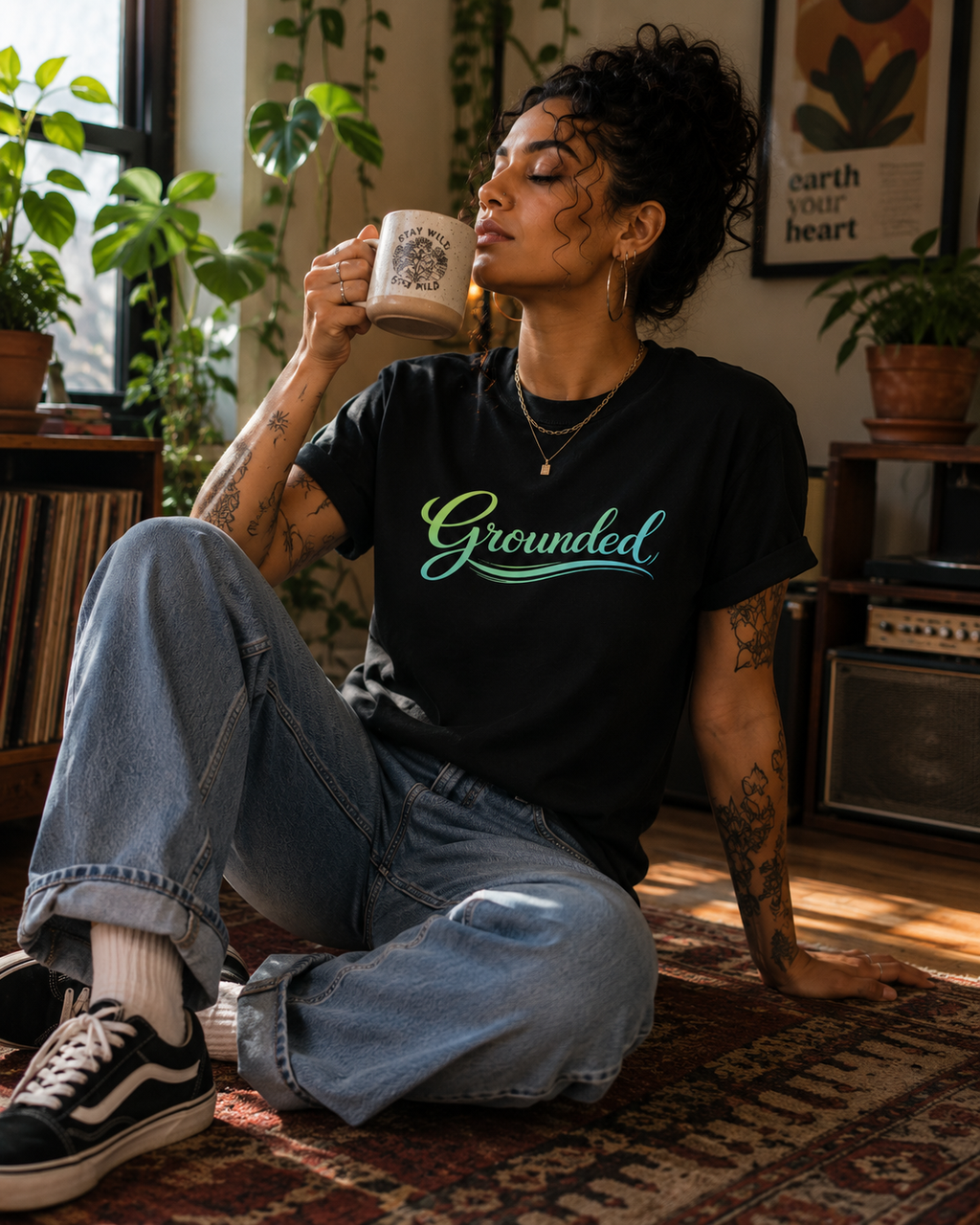 A woman sits on a patterned rug in a cozy room, holding a mug and wearing a black graphic t-shirt with the word 'Grounded' in green and blue script. She is wearing light-wash blue jeans and black sneakers.