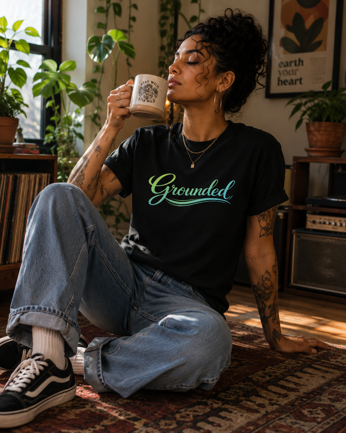 A woman sits on a patterned rug in a cozy room, holding a mug and wearing a black graphic t-shirt with the word 'Grounded' in green and blue script. She is wearing light-wash blue jeans and black sneakers.