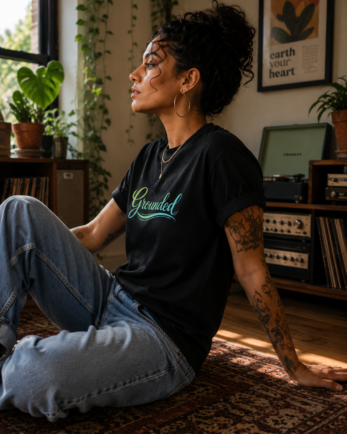 A woman sitting on a patterned rug in a cozy room, wearing a black graphic t-shirt with the word 'Grounded' in a neon green and blue script font, paired with light blue jeans.