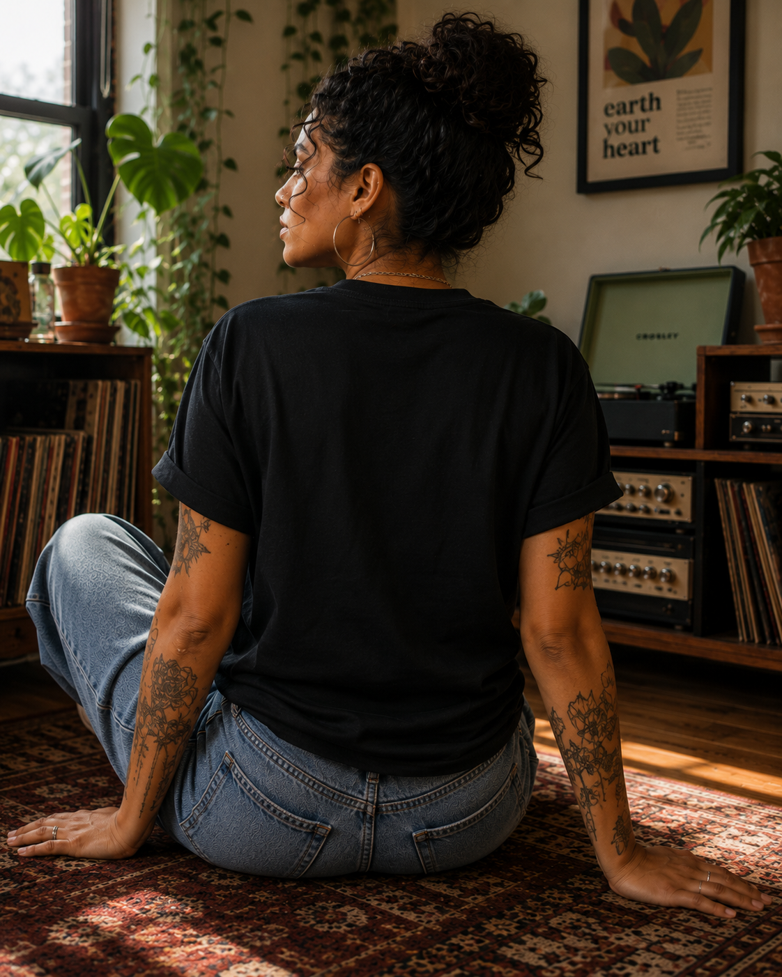 A woman with curly dark hair sits cross-legged on a patterned rug in a cozy room, viewed from behind. She is wearing a plain black short-sleeve t-shirt and light blue jeans.