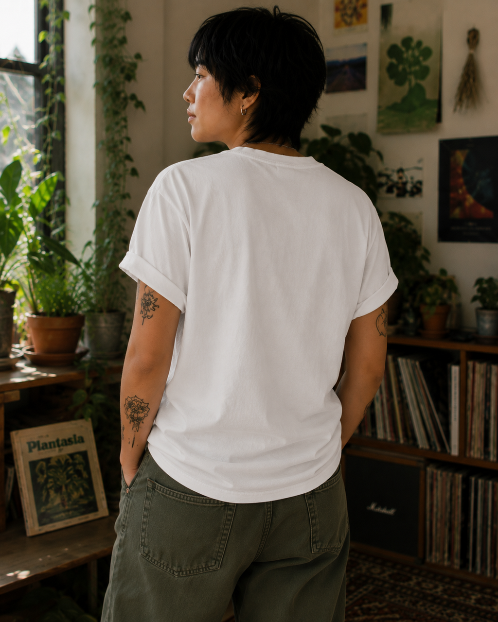A person with short dark hair stands with their back to the camera wearing a plain white short-sleeve t-shirt and olive green pants in a cozy, plant-filled room.
