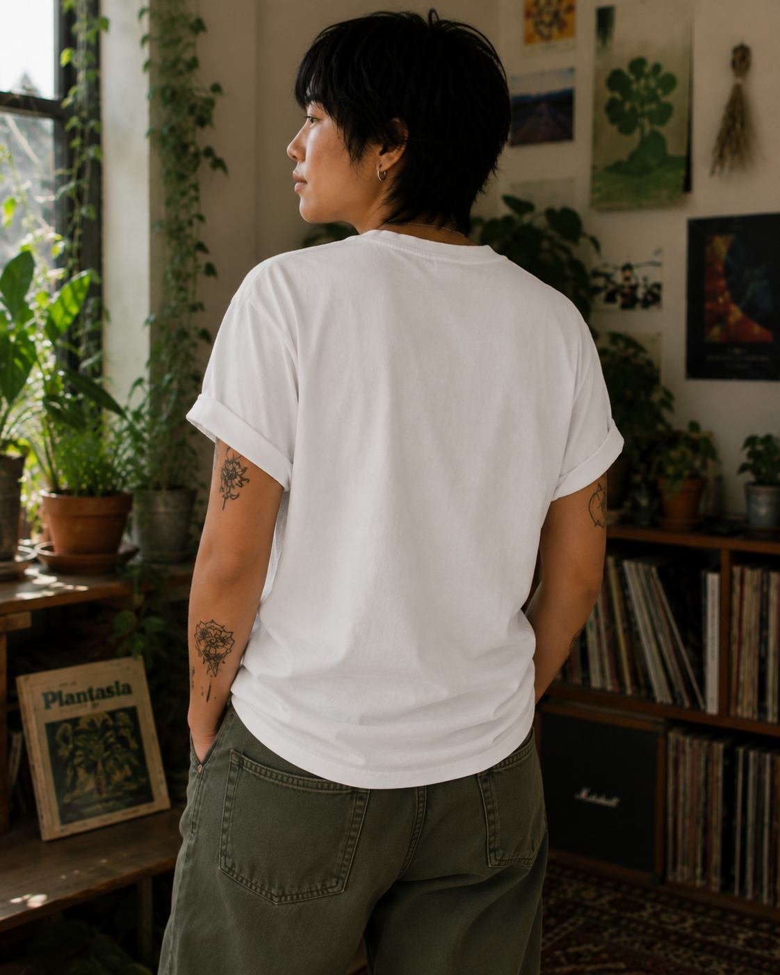 A person with short dark hair stands with their back to the camera wearing a plain white short-sleeve t-shirt and olive green pants in a cozy, plant-filled room.