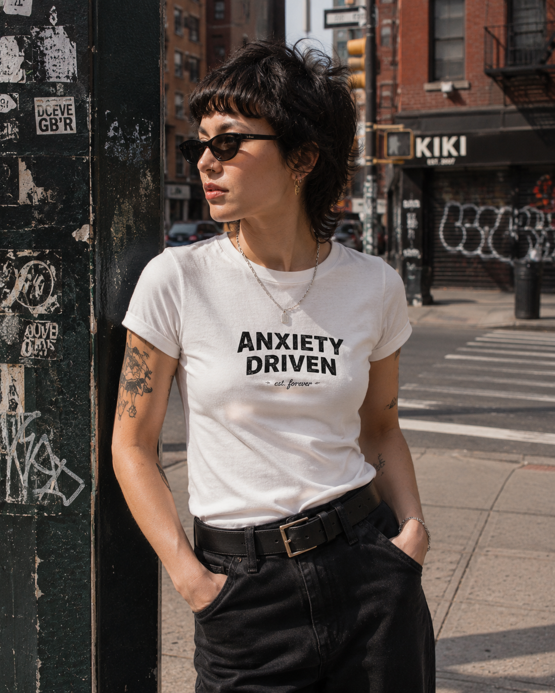 A woman wearing a white graphic t-shirt with the text 'ANXIETY DRIVEN' printed in black, standing on a city street next to a graffiti-covered pole.