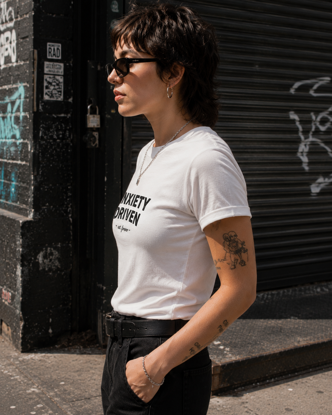A woman wearing a white crewneck t-shirt with black text that reads 'SOXITY IS DRIVEN' stands in profile against a black metal shutter storefront.