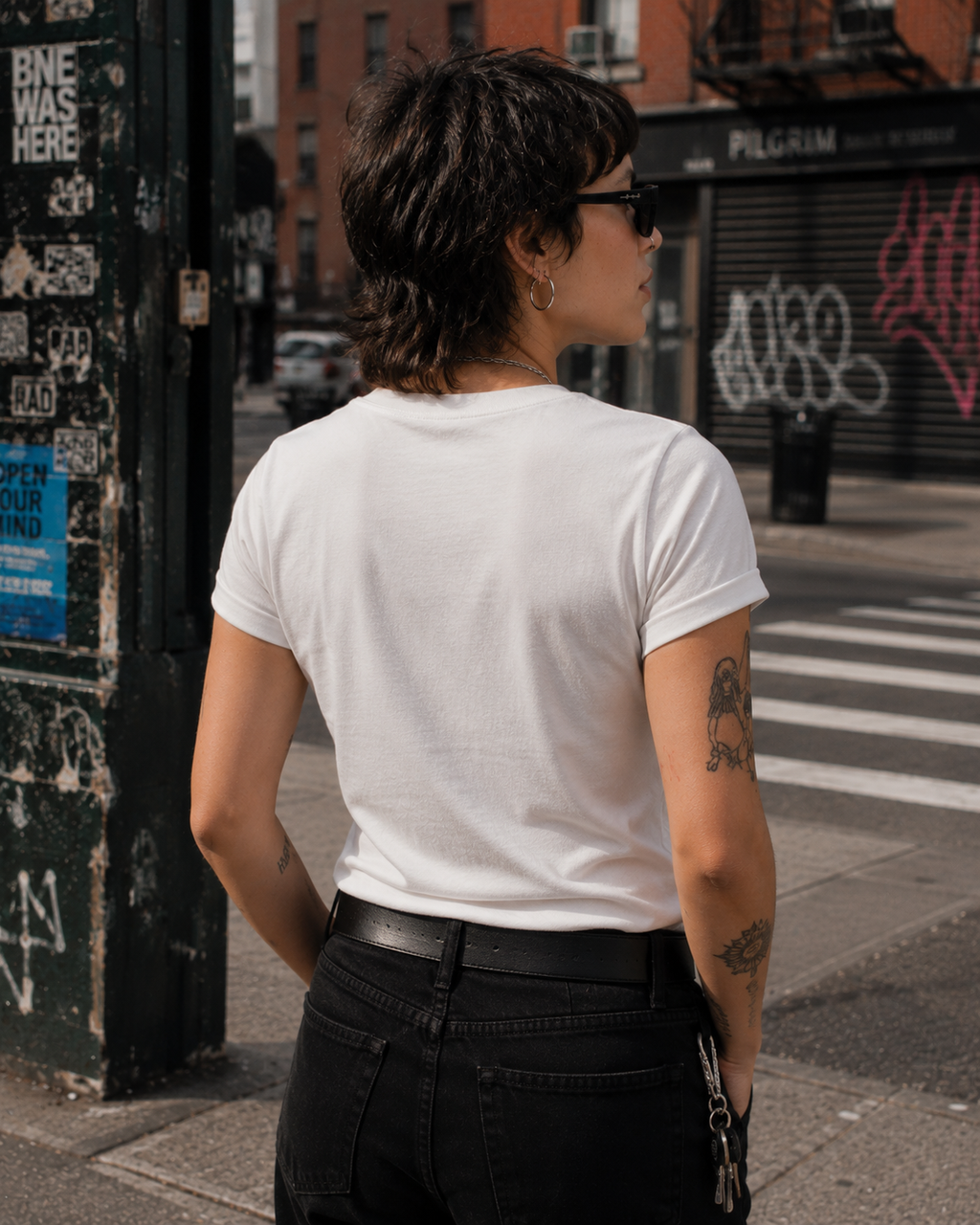 A person standing on a city street, shown from the back, wearing a plain white short-sleeve T-shirt tucked into black jeans.
