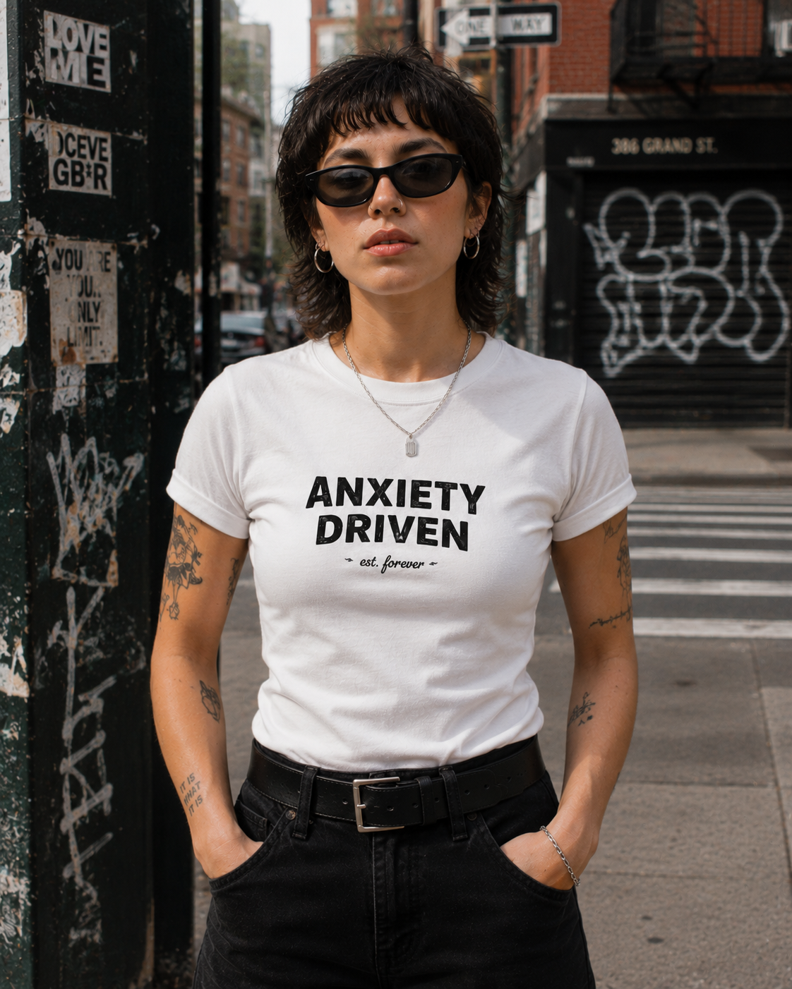 A woman wearing a white graphic t-shirt with the text 'ANXIETY DRIVEN' and 'all forward' in black, standing on a city street.