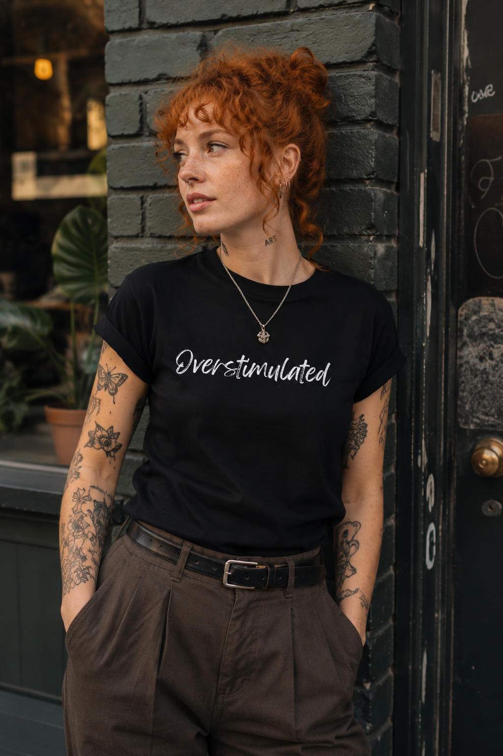 A woman with curly red hair and tattoos leans against a dark brick wall wearing a black t-shirt with the white script text 'Over-stimulated' and brown pleated trousers.
