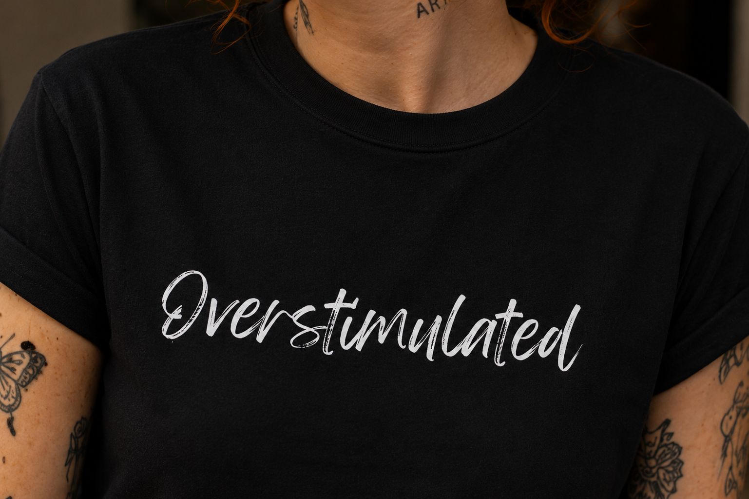 A close-up of a person wearing a black short-sleeve crewneck t-shirt with the word 'Overstimulated' printed across the chest in a white cursive script.