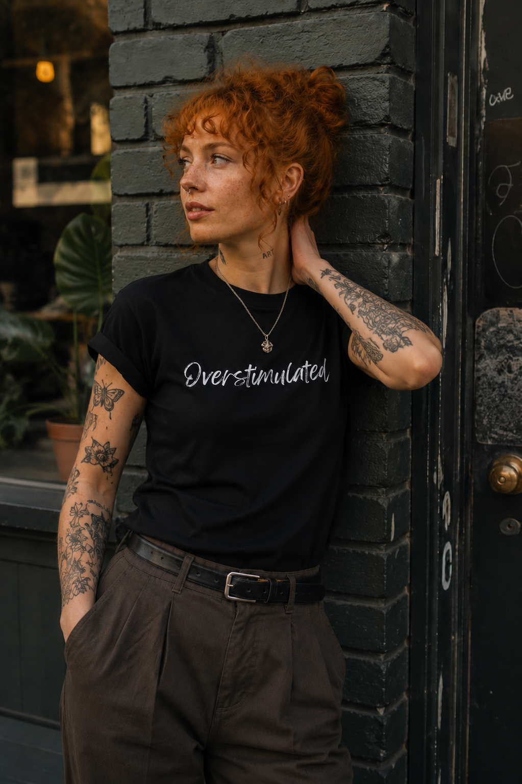 A woman with curly red hair and tattoos leans against a dark brick wall wearing a black t-shirt with the word 'Overstimulated' printed in white script, paired with brown pants.