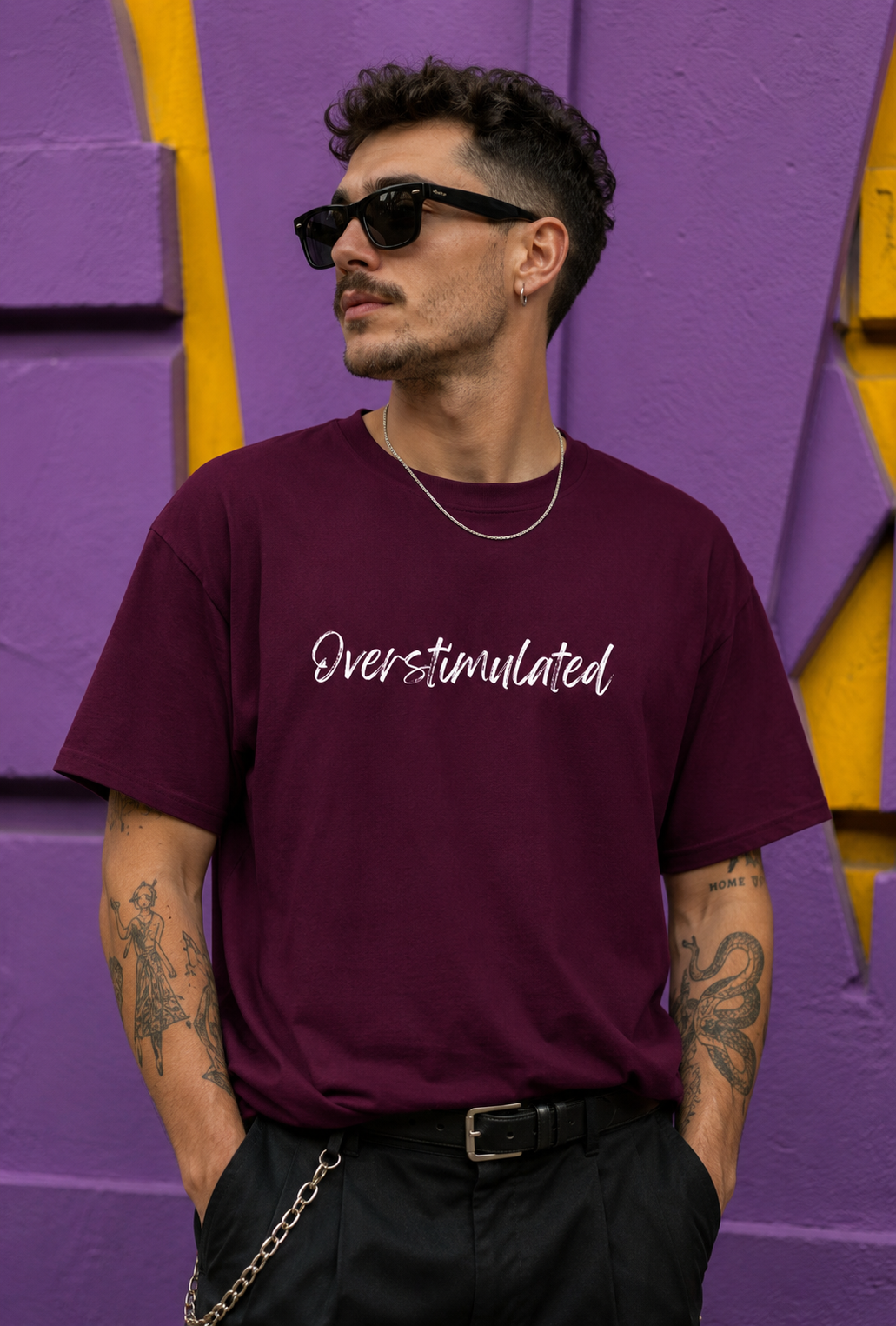 A man wearing a maroon crewneck t-shirt with 'Overstimulated' printed in white script across the chest, paired with black pants, sunglasses, and a silver chain necklace, standing against a purple and yellow textured wall.