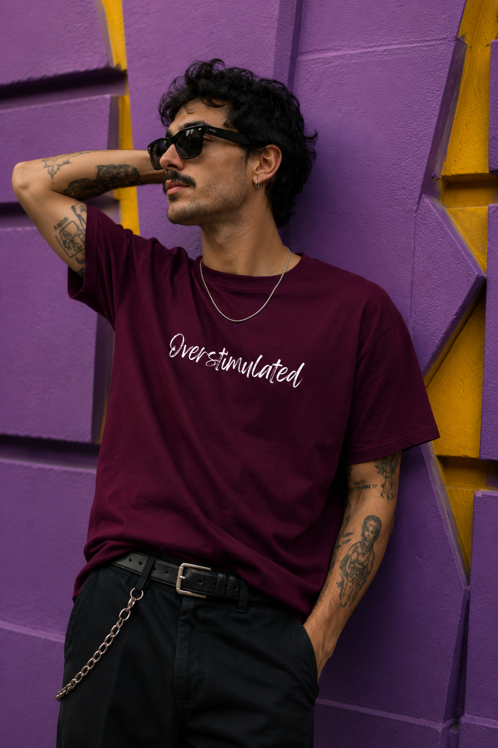 A man wearing a maroon t-shirt with 'Overstimulated' printed in white script, paired with black pants and sunglasses, posing against a purple and yellow geometric wall.