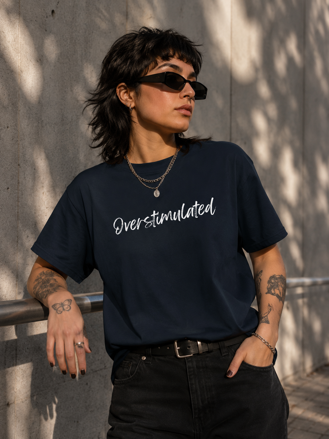 A person wearing a navy blue graphic t-shirt with the word 'Overstimulated' printed in white script, paired with black jeans and sunglasses, standing against a concrete wall with dappled sunlight.