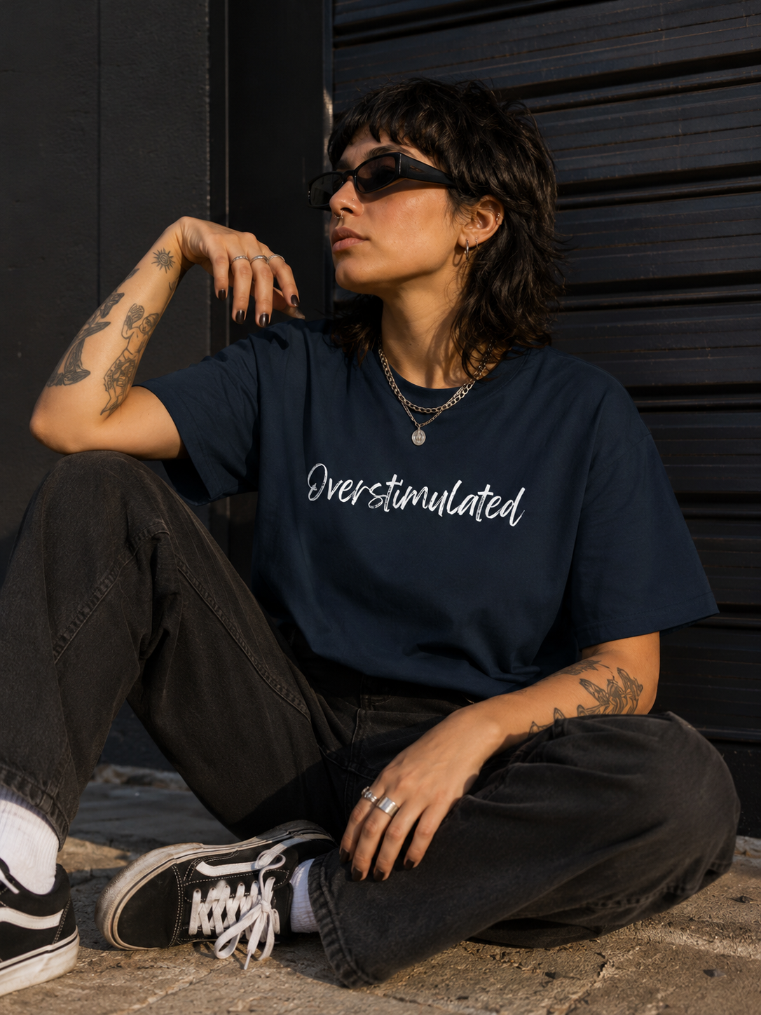 A person sitting outdoors against a dark metal wall, wearing a navy blue graphic T-shirt with the word 'Overstimulated' in white script, black jeans, black sneakers, and sunglasses.